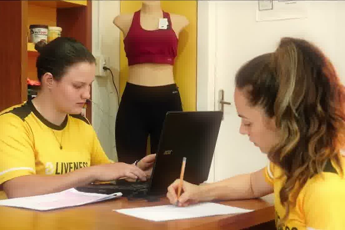 Duas colaboradoras de camiseta Liveness trabalhando na recepção de uma unidade — uma no notebook, outra preenchendo uma ficha sobre a mesa